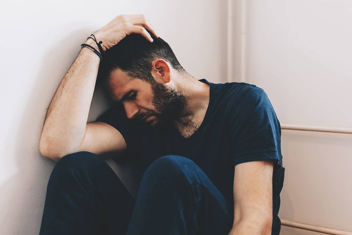 Bearded man in blue shirt sitting against wall, head down with hand on head, depicting depression and emotional distress associated with addiction