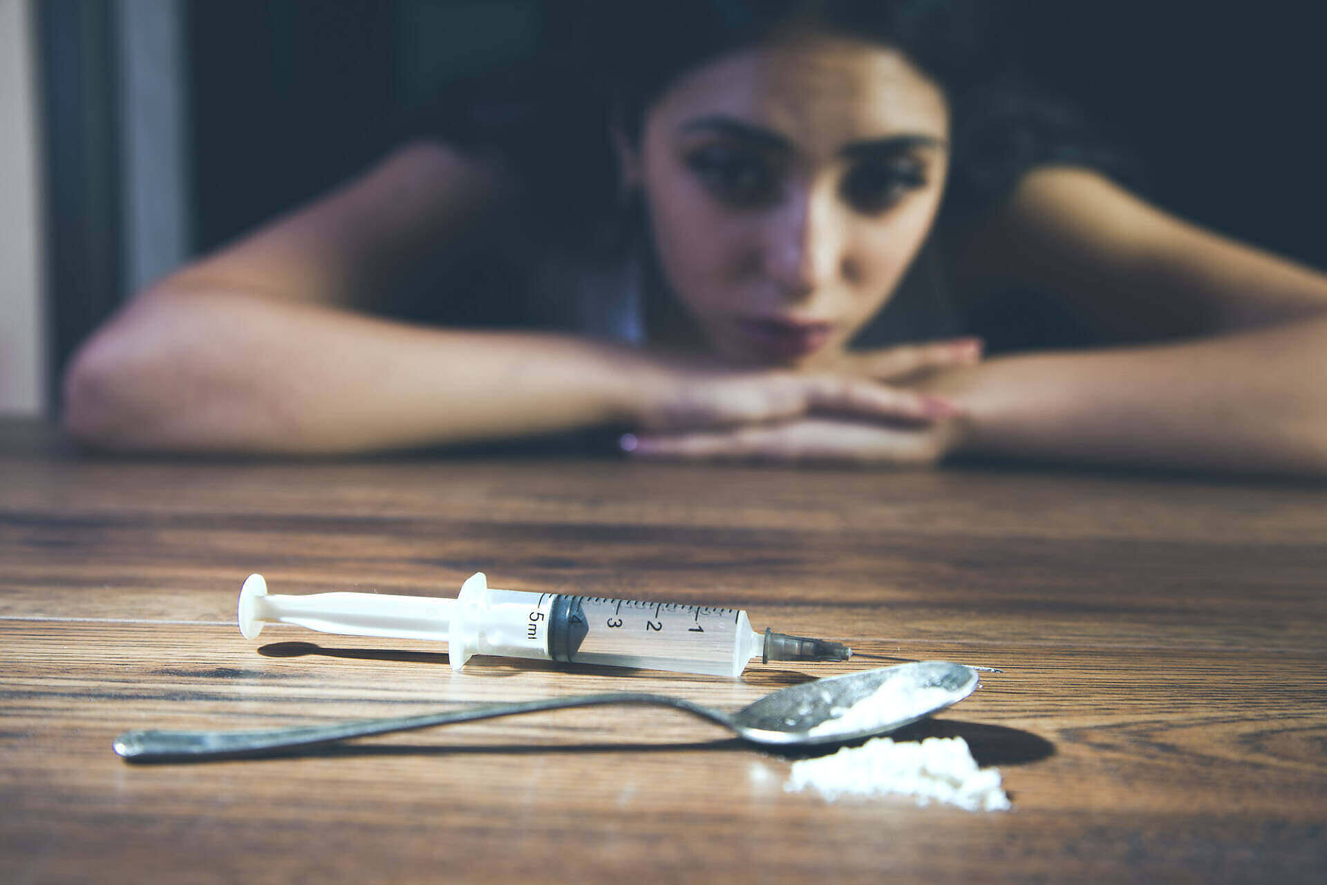 Woman looking distressed with drug paraphernalia including syringe and powder on table, depicting struggles with addiction