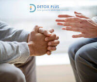 Close-up of two people's hands during a counseling session, with one person clasping their hands and the other gesturing supportively. Detox Plus UK logo and tagline "Your guide to addiction recovery" visible in the top left corner.