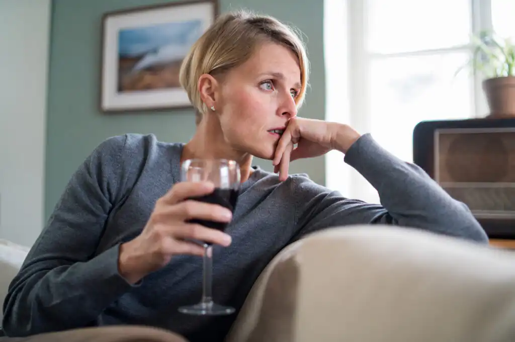 Concerned woman holding glass of red wine, looking pensive in home setting, possible signs of alcohol dependency