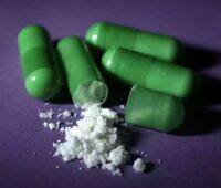 Several green drug capsules next to a small pile of white powdery substance on a purple background