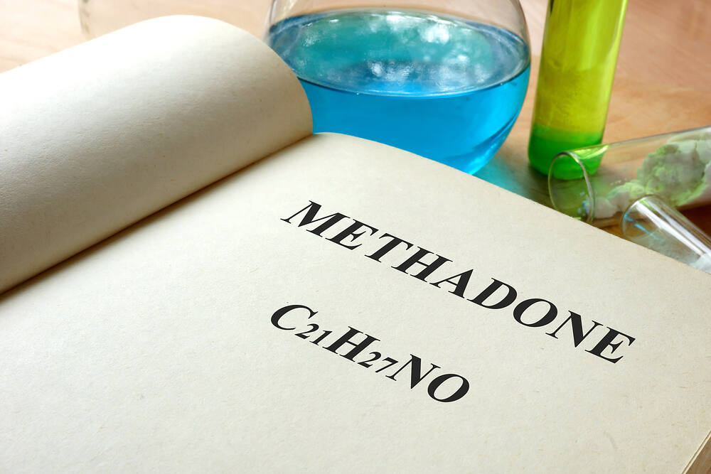 Open book displaying the word 'Methadone' and its chemical formula C21H27NO, with laboratory glassware in the background