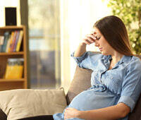 Pregnant woman sitting on a couch looking distressed, holding her head with one hand and her belly with the other, illustrating the struggle with addiction during pregnancy