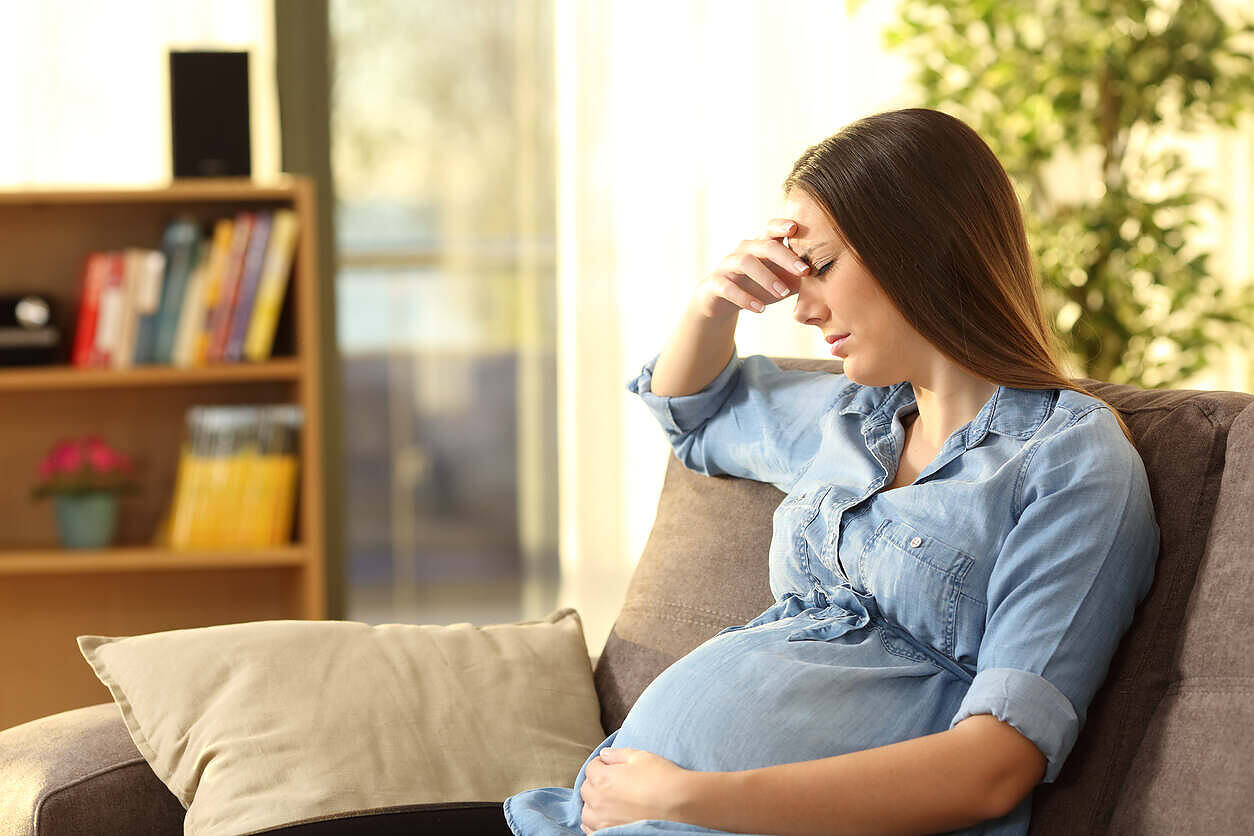 Pregnant woman sitting on a couch looking distressed, holding her head with one hand and her belly with the other, illustrating the struggle with addiction during pregnancy