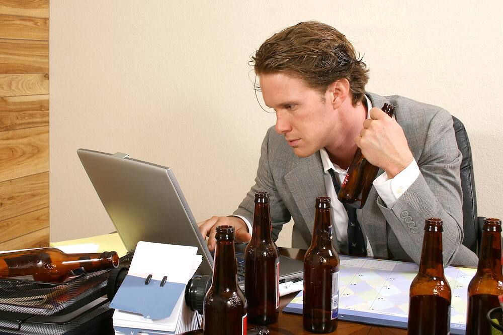 Man in suit working on laptop surrounded by beer bottles, depicting a functioning alcoholic in an office setting