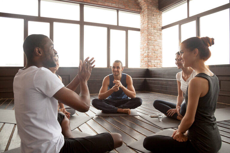 Diverse group of people sitting in a circle on yoga mats during a therapy or support group session, with a man gesturing as he speaks to the others in a bright, modern room with exposed brick walls