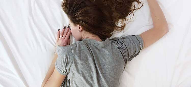 Woman in gray shirt lying face down on white bedding, illustrating sleep difficulties associated with addiction