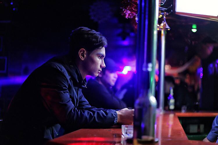 Young man sitting alone at a bar in a dimly lit nightclub, surrounded by alcohol bottles, illustrating potential student substance abuse issues