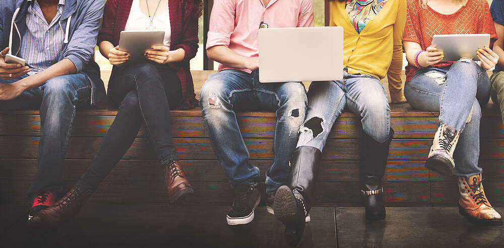 Group of diverse young adults sitting together, using tablets and smartphones, highlighting digital connectivity and potential substance abuse risks