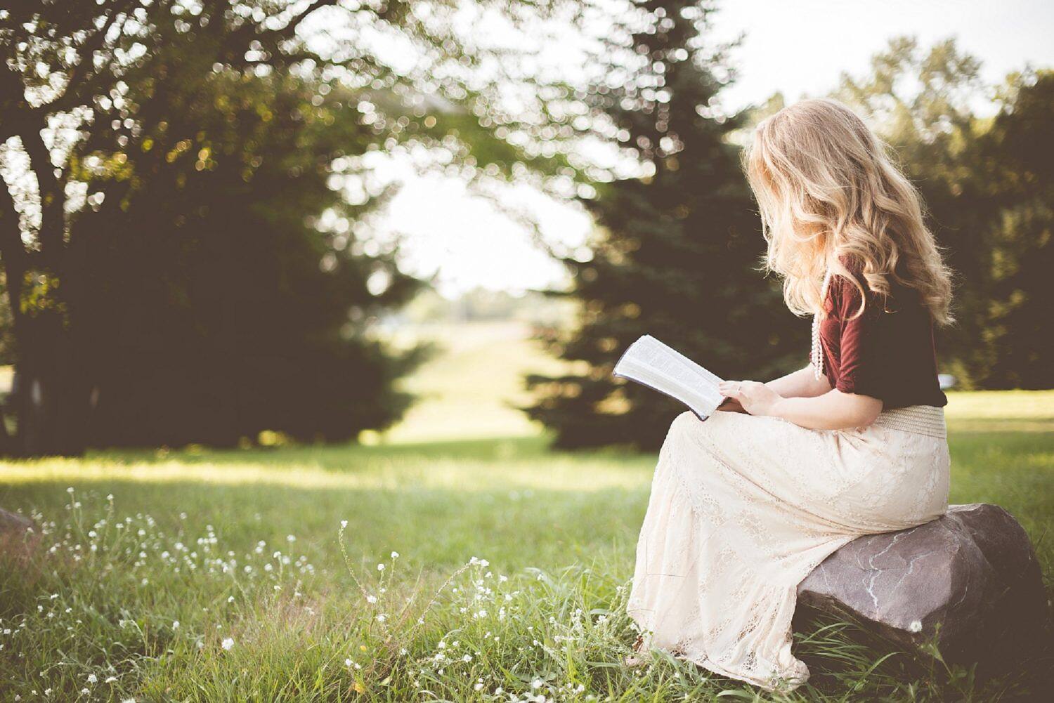 Woman with blonde hair sitting on a rock in a peaceful garden, reading a book, symbolizing tranquility and self-reflection in addiction recovery