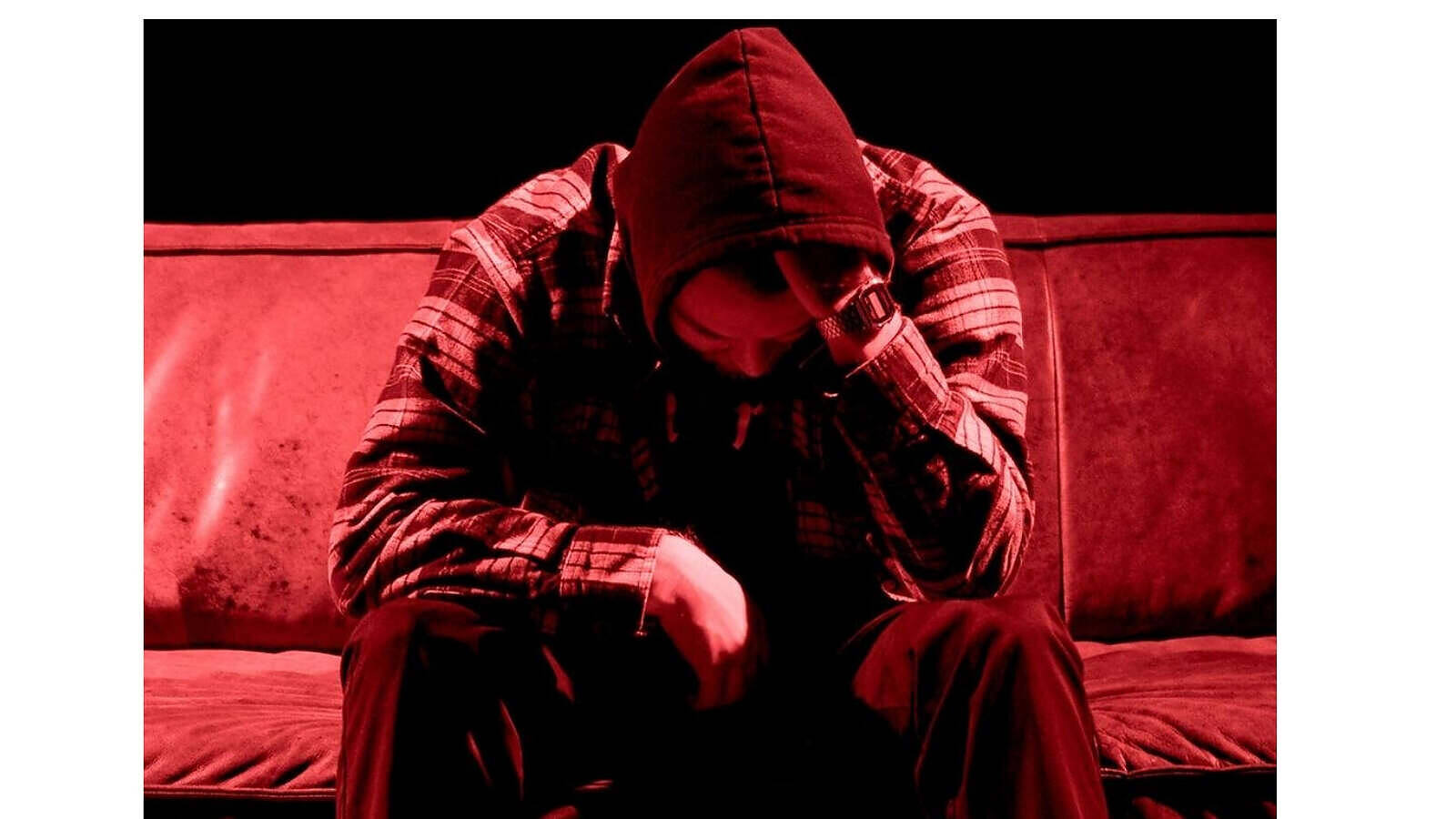 Despondent figure in red hoodie and plaid shirt sitting hunched over on a couch, symbolizing the isolation and despair often associated with substance abuse