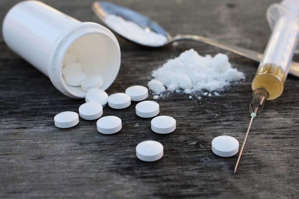 Opiate addiction paraphernalia including white pills spilled from a bottle, powdered substance, spoon, and syringe on a dark wooden surface