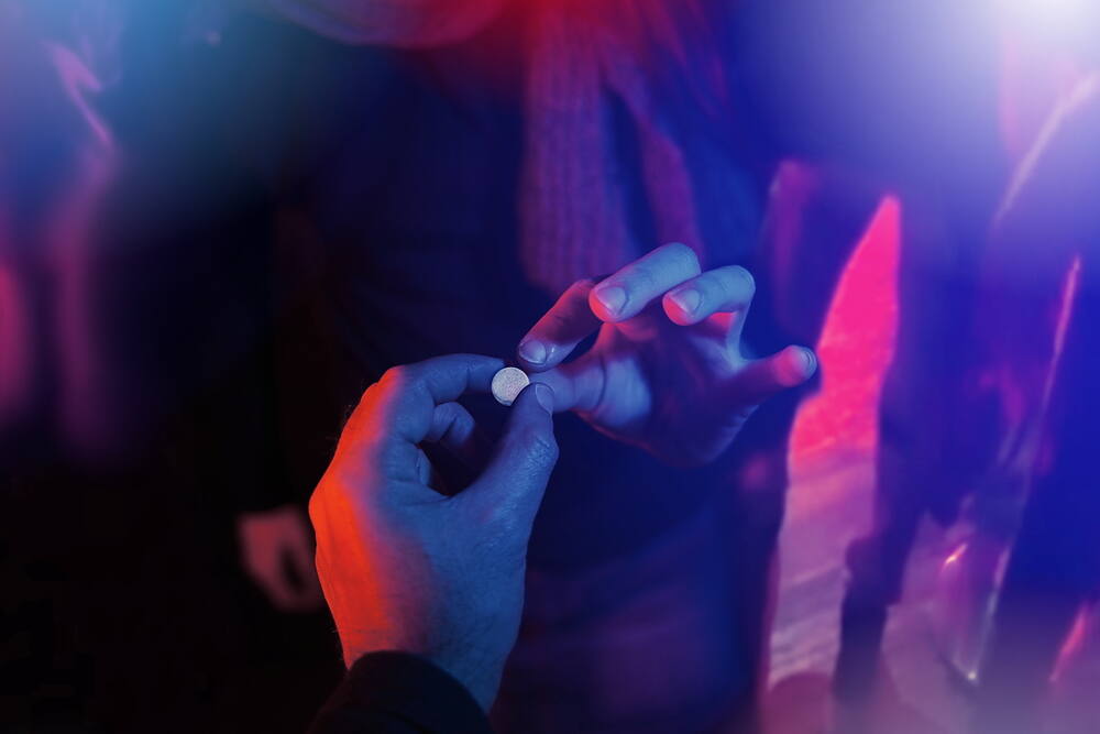 Close-up of a hand holding an ecstasy pill in a dark nightclub environment, illuminated by blue and red lights, highlighting the risks of drug use in party scenes