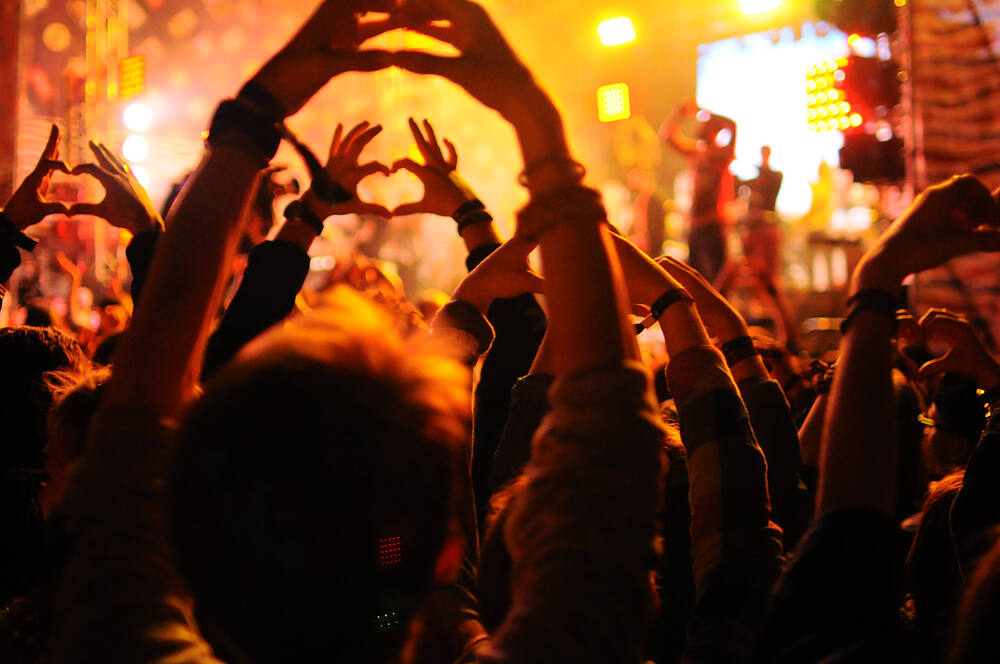 Crowd at a nighttime music festival or rave, with silhouettes of people making heart shapes with their hands against a backdrop of bright orange stage lights, illustrating the environment where ecstasy use often occurs