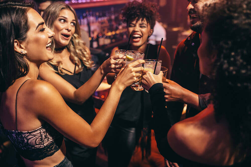 Group of young adults laughing and toasting with cocktails at a crowded bar, illustrating social drinking behavior