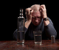 Distressed elderly man with hands on head sitting at table with bottle of alcohol and glass, illustrating the struggle with alcohol addiction in later life