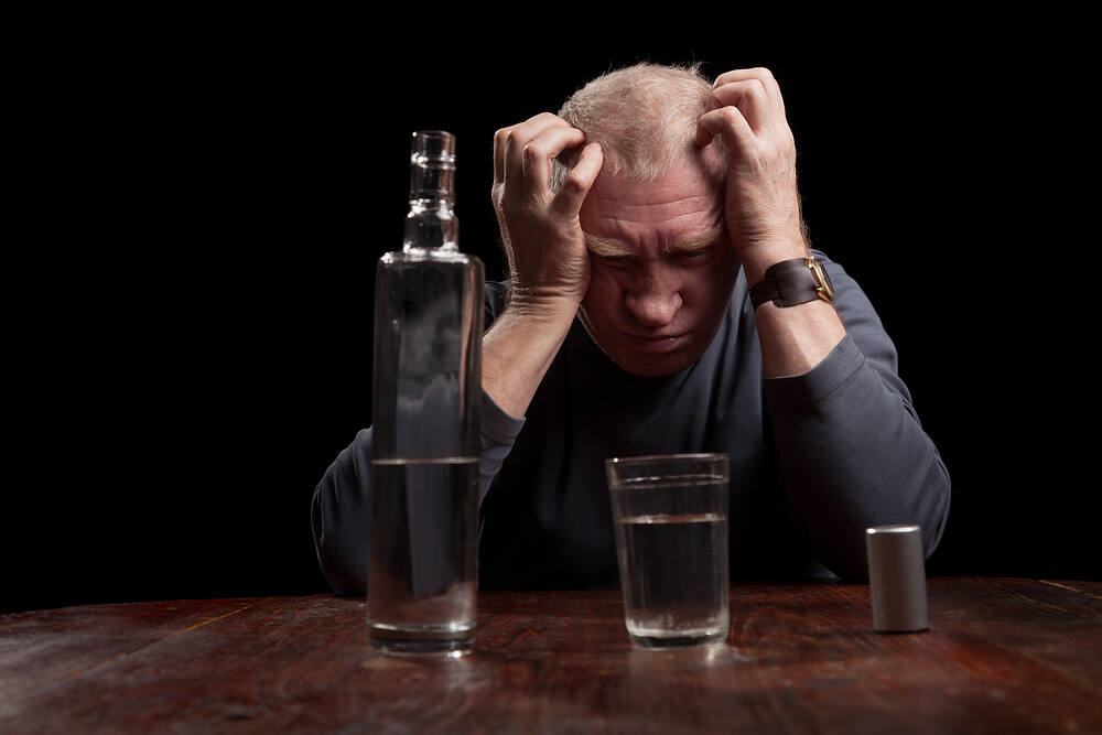 Distressed elderly man with hands on head sitting at table with bottle of alcohol and glass, illustrating the struggle with alcohol addiction in later life