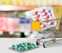 Miniature shopping cart filled with blister packs of various prescription medications, including red and white capsules, alongside a credit card, symbolizing the ease of access to potentially addictive drugs through online pharmacies