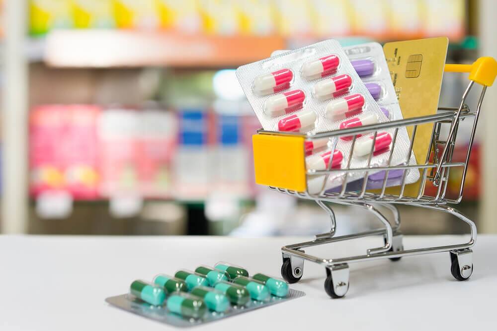 Miniature shopping cart filled with blister packs of various prescription medications, including red and white capsules, alongside a credit card, symbolizing the ease of access to potentially addictive drugs through online pharmacies