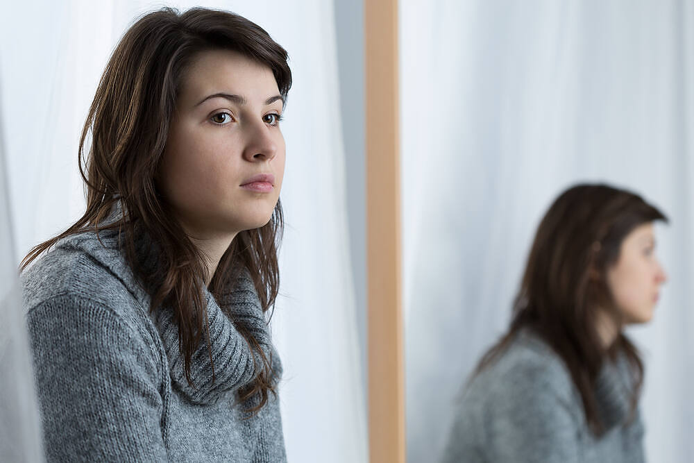 Young woman in gray sweater with thoughtful expression, looking away from mirror reflection