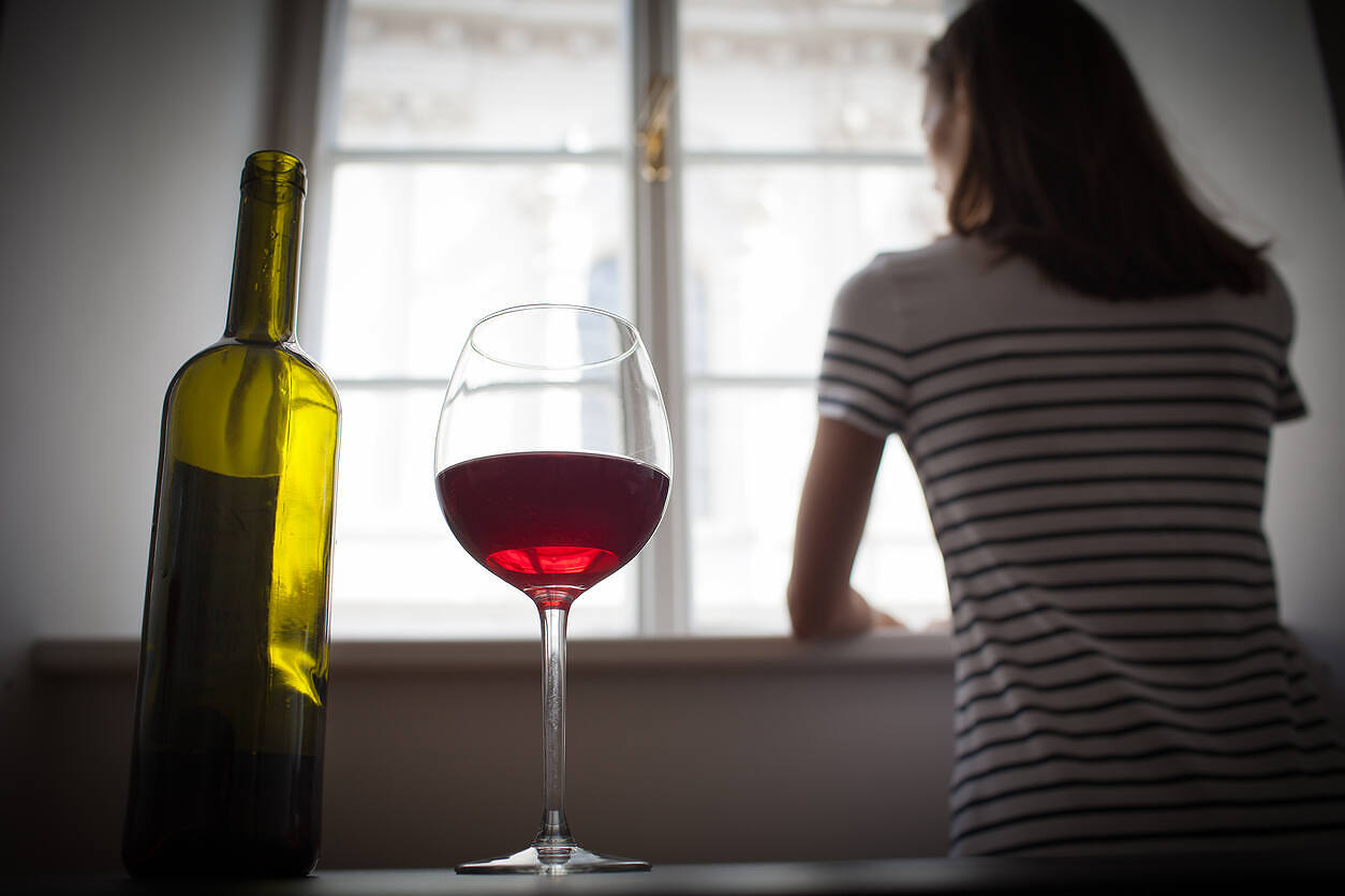 Woman looking out window with wine bottle and glass in foreground, depicting alcohol addiction and isolation