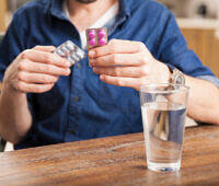 Man in blue shirt holding medication blister packs, with a glass of water on a wooden table, illustrating gabapentin use and potential for misuse