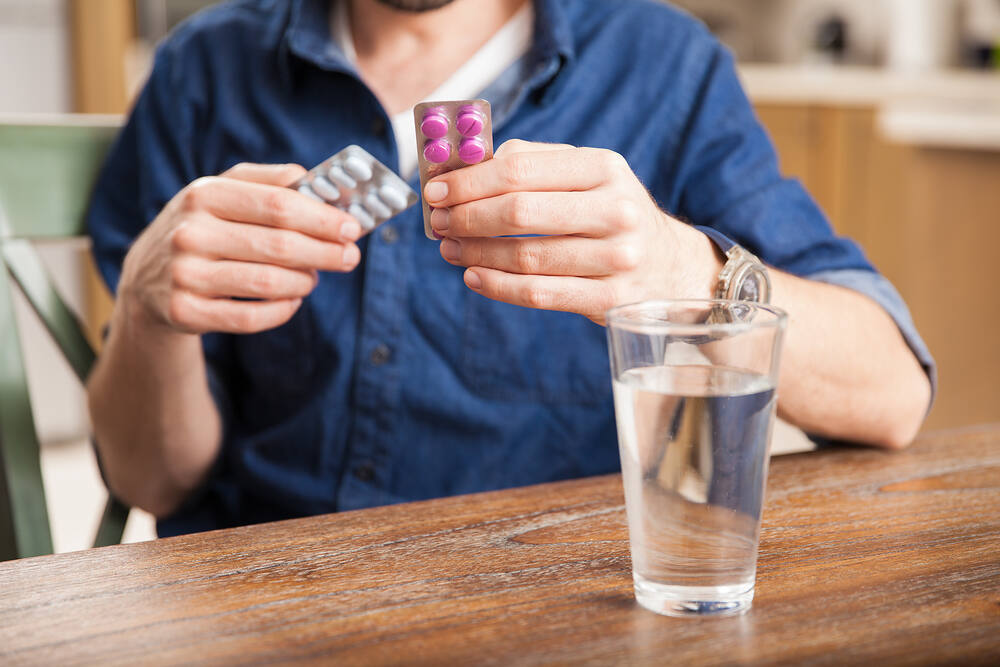 Man in blue shirt holding medication blister packs, with a glass of water on a wooden table, illustrating gabapentin use and potential for misuse