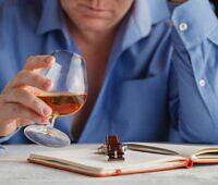 Person in blue shirt holding a glass of alcohol next to an open notebook, depicting work-related stress and alcohol use