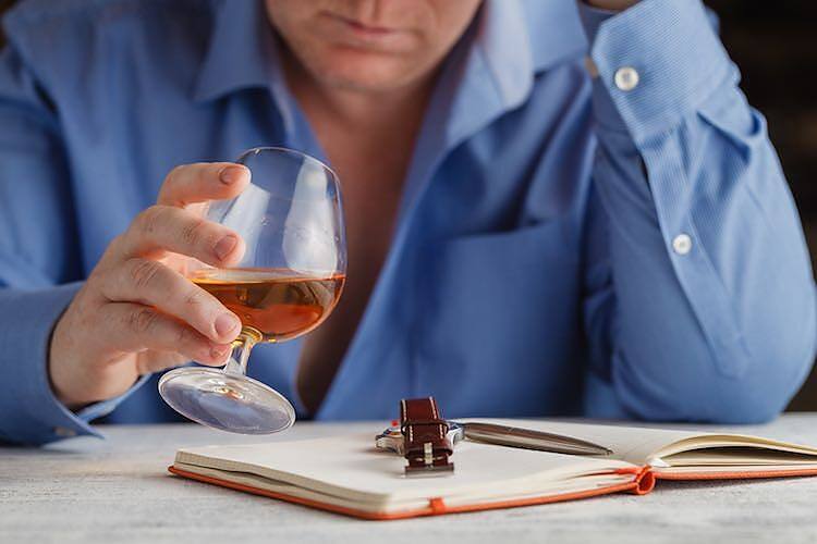 Person in blue shirt holding a glass of alcohol next to an open notebook, depicting work-related stress and alcohol use