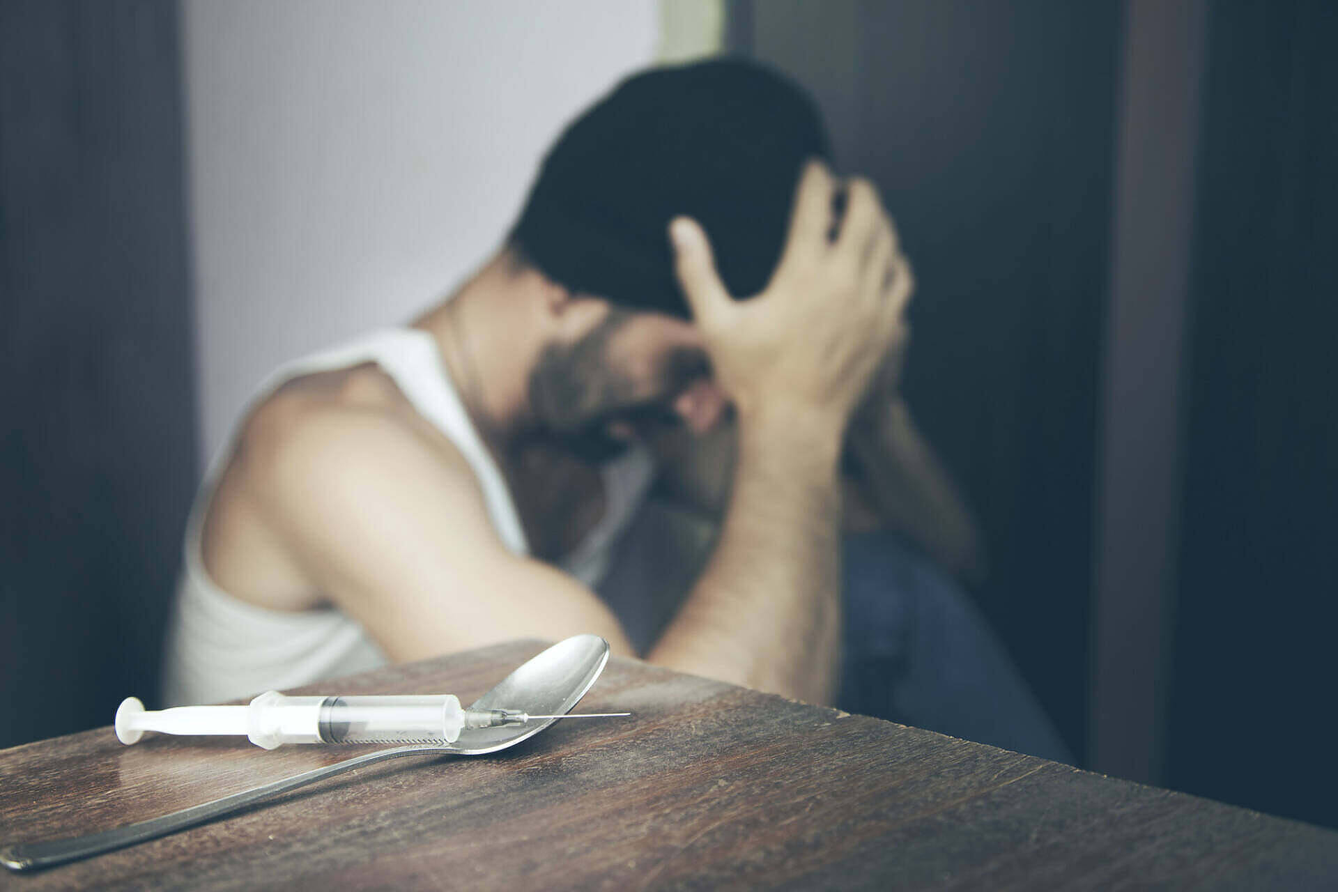 Distressed man with head in hands sitting behind drug paraphernalia including syringe and spoon, illustrating the struggle with heroin addiction