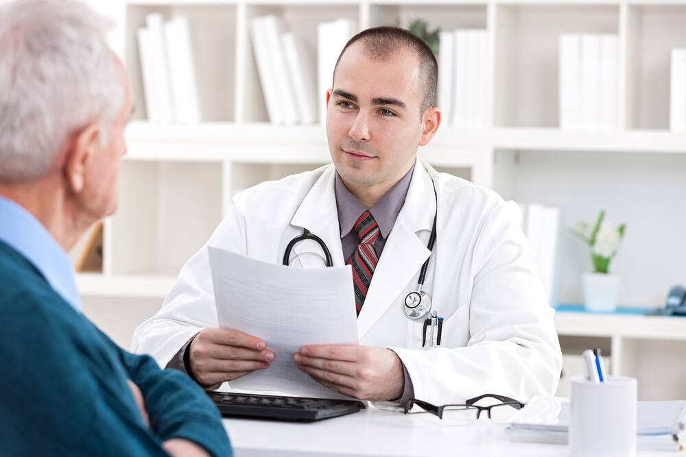 Young male doctor in white coat reviewing medical documents with elderly male patient during consultation