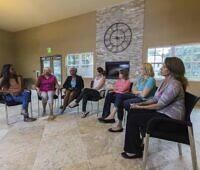 Diverse group of women participating in a therapy session at a rehabilitation centre, sitting in a circle in a modern, well-lit room with a large clock on the wall.