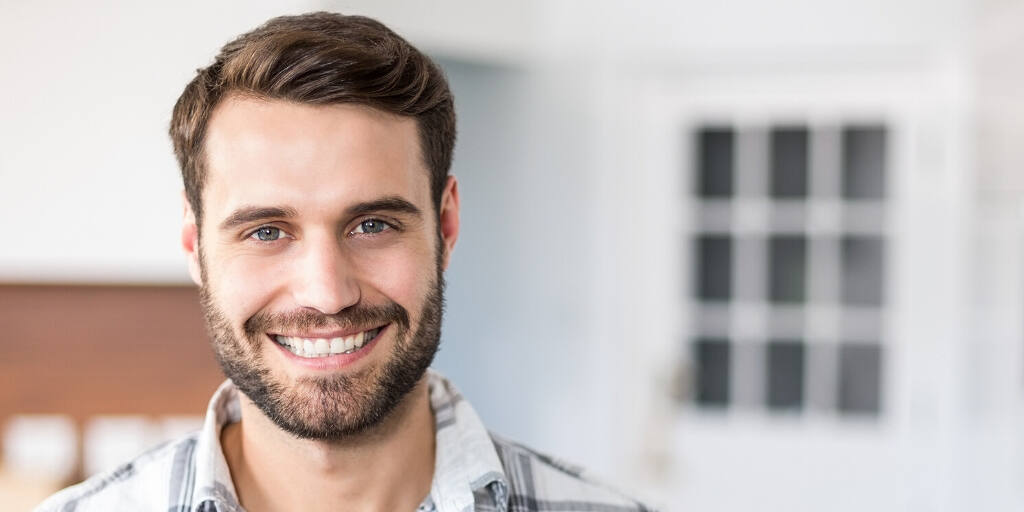 Headshot of a smiling bearded man with brown hair in a casual shirt, standing in a bright office space