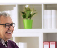 Smiling elderly man in glasses speaking with a young doctor in a bright, modern medical office with bookshelves