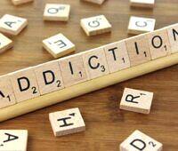 The word 'ADDICTION' spelled out using Scrabble tiles on a wooden tile rack, with scattered Scrabble tiles in the background.