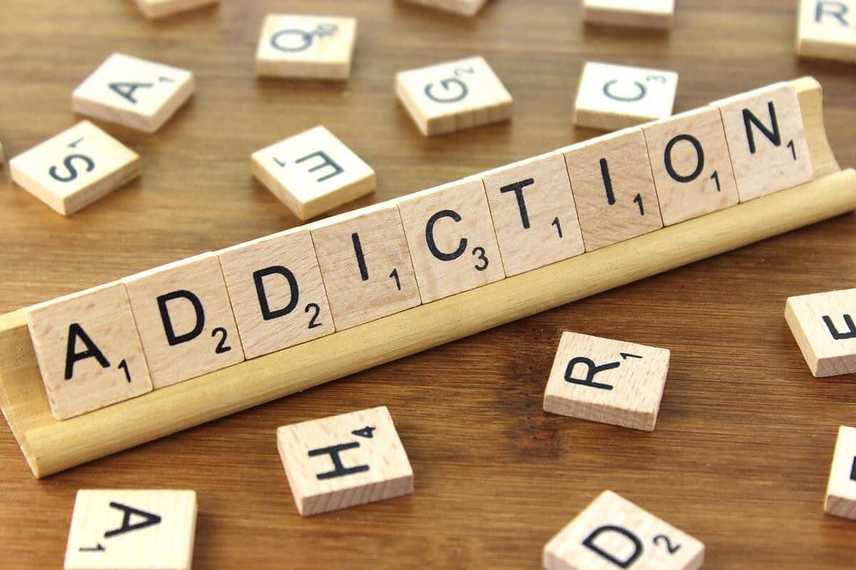 The word 'ADDICTION' spelled out using Scrabble tiles on a wooden tile rack, with scattered Scrabble tiles in the background.