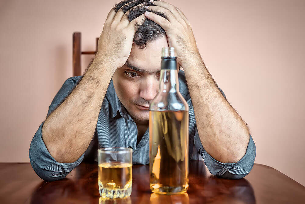 Distressed man holding his head with bottle and glass of alcohol on table, depicting alcohol addiction and anxiety