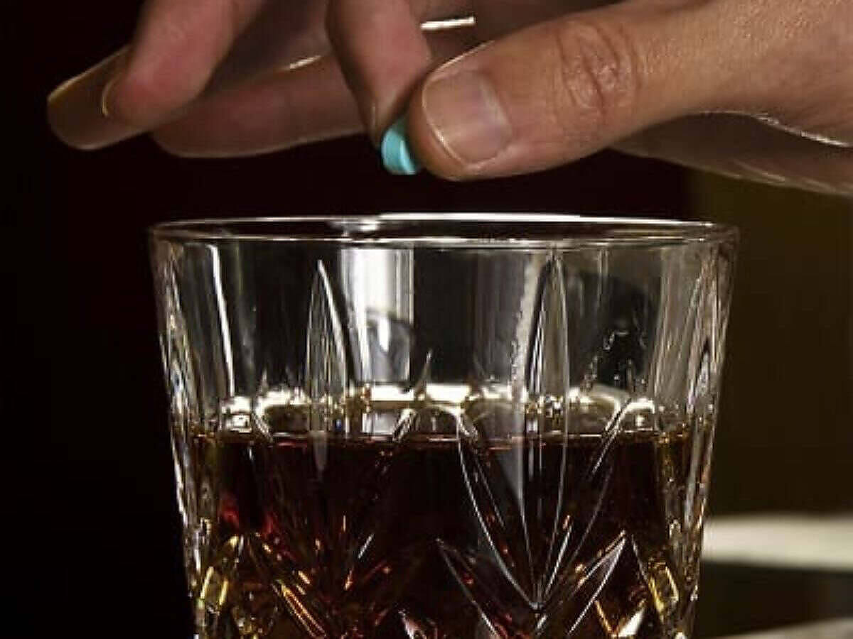 Hand dropping a pill, possibly Rohypnol, into a glass of dark liquid, likely alcohol, illustrating the dangerous act of drink spiking