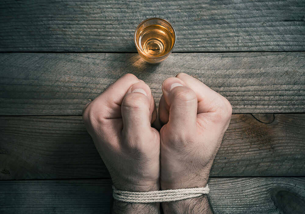Clenched fists tied with rope next to a glass of alcohol on a wooden surface, symbolizing addiction struggles