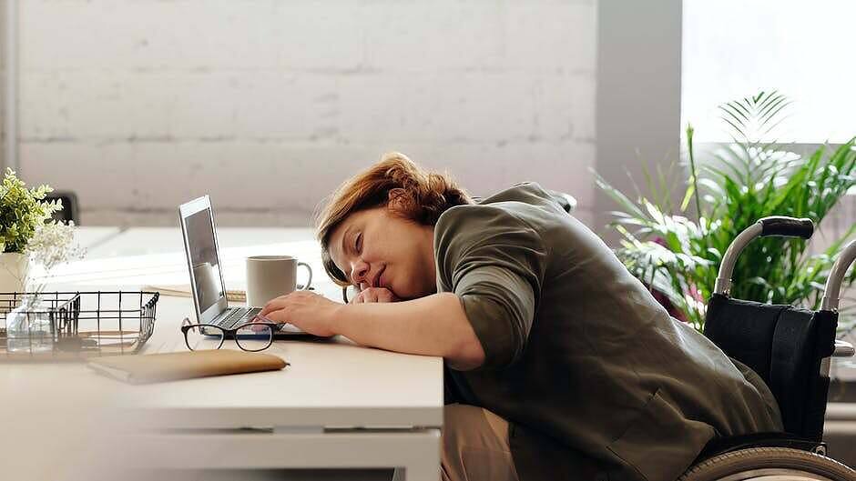 Woman in wheelchair sleeping at desk with laptop, showing signs of extreme fatigue possibly related to substance use