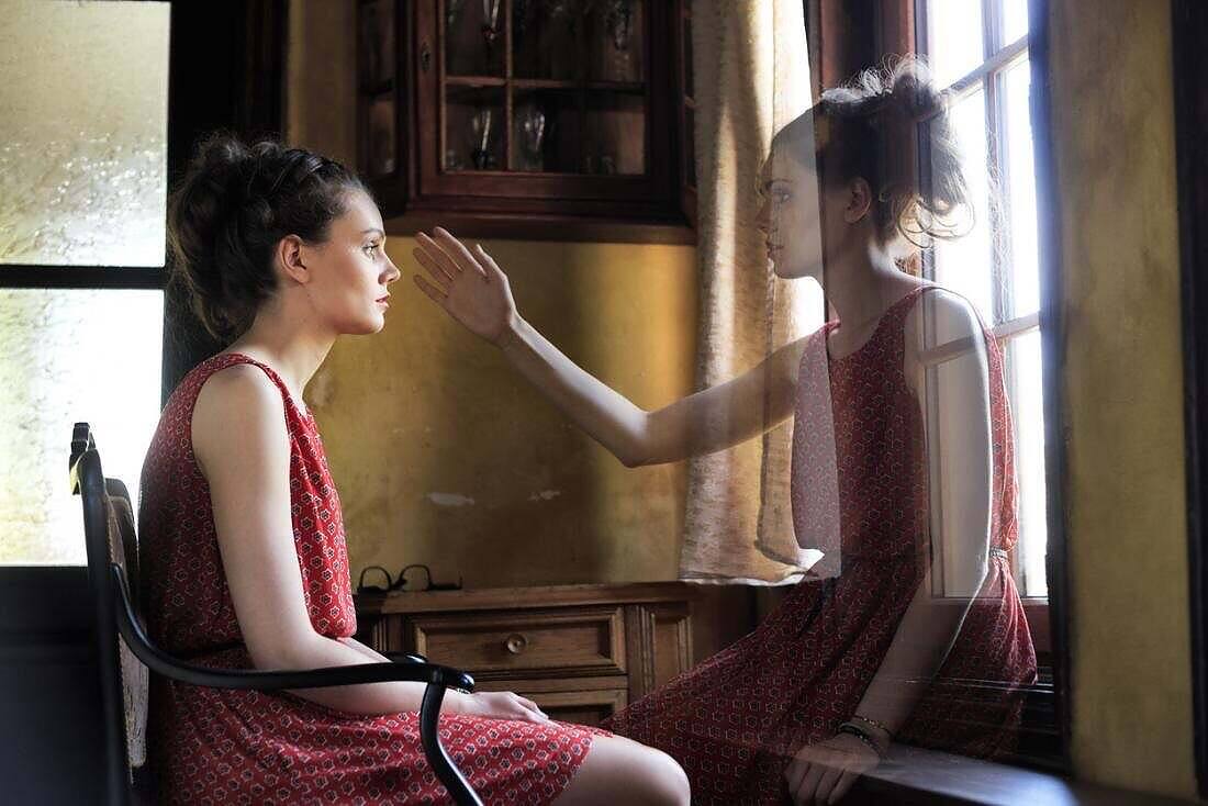 Woman in red dress sitting by window, with a semi-transparent version of herself reaching back to her, symbolizing confrontation with past or future self during recovery