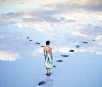 Woman in tie-dye dress walking on stepping stones across reflective water surface mirroring sky and clouds, symbolizing the path to healing and sobriety