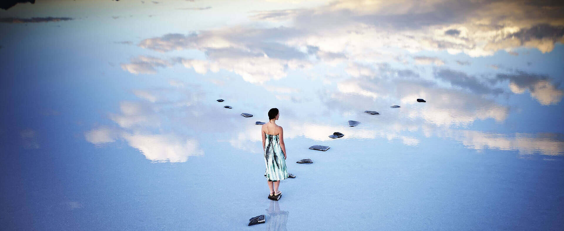 Woman in tie-dye dress walking on stepping stones across reflective water surface mirroring sky and clouds, symbolizing the path to healing and sobriety