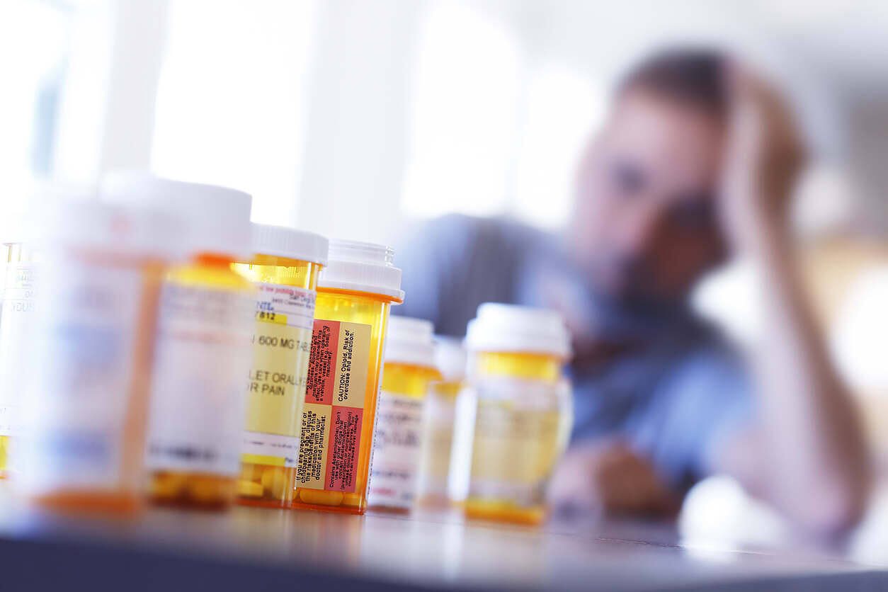 Multiple prescription medication bottles in foreground with blurred image of distressed person holding head in background, illustrating struggle with prescription drug dependence