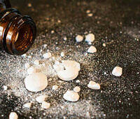 Spilled white pills and powder next to an open brown pill bottle on dark surface, illustrating drug addiction dangers