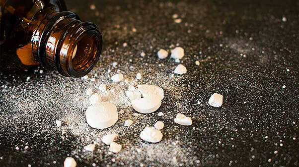 Spilled white pills and powder next to an open brown pill bottle on dark surface, illustrating drug addiction dangers