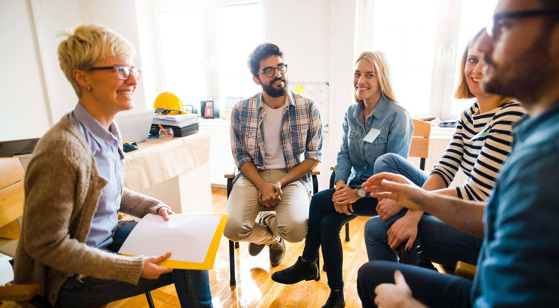 Diverse group of people sitting in a circle during a therapy session at an addiction treatment center, led by a female therapist