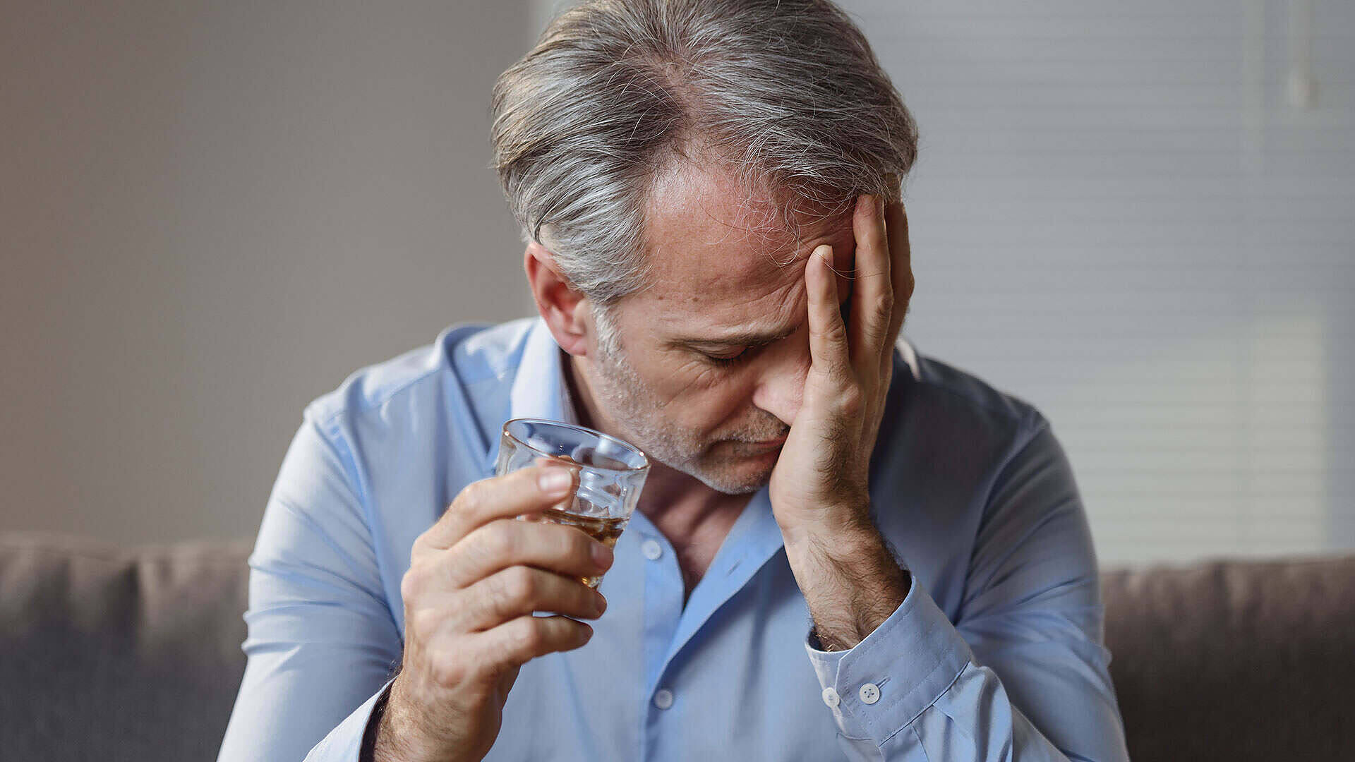 Middle-aged man with grey hair holding a glass of alcohol, looking distressed and covering his face with his hand, illustrating alcohol dependency