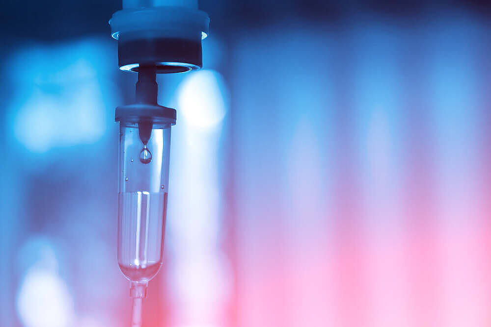 Close-up of an IV drip with a clear liquid, possibly glutathione, against a blue and pink blurred background in a medical setting