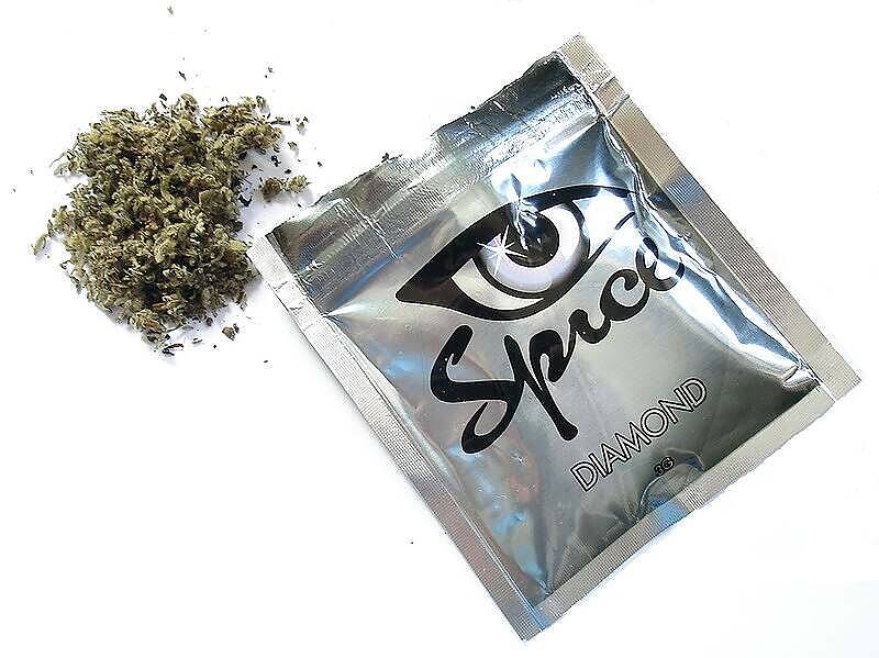 Packet of synthetic cannabis labeled "Spice Diamond" with eye logo, next to pile of dried herbal material, illustrating the appearance of dangerous synthetic drugs